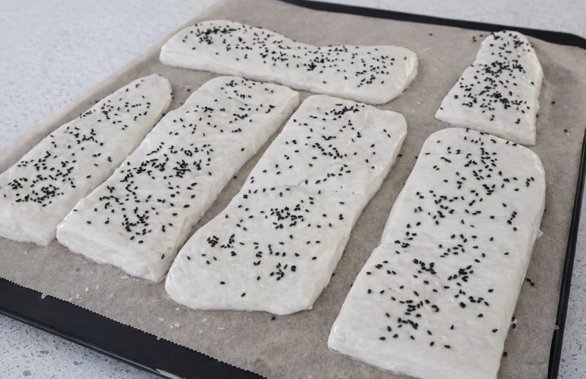 Rå ciabattabrød toppet med svarte frø og klare for steking på et stekebrett med bakepapir.