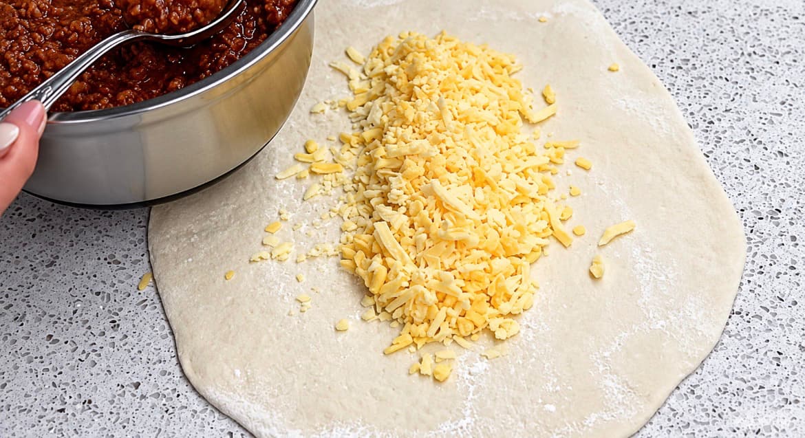 Revet ost legges midt på den utkjevlede pizzadeigen før kjøttsausen tilsettes.