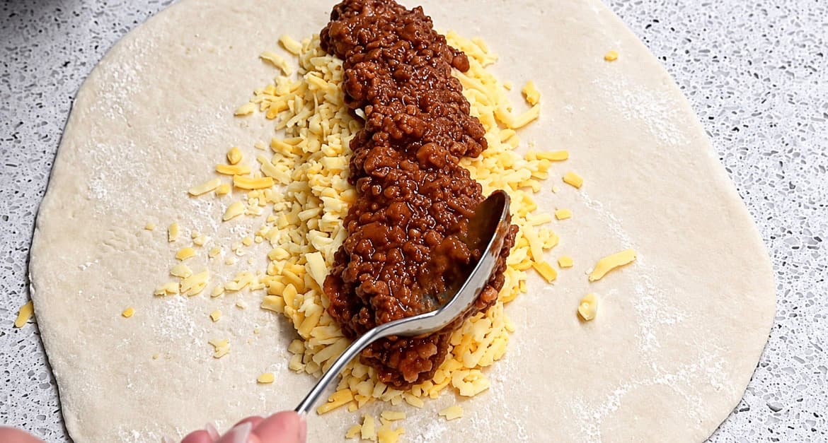 Kjøttsaus legges over revet ost midt på den utkjevlede deigen til innbakt pizza.