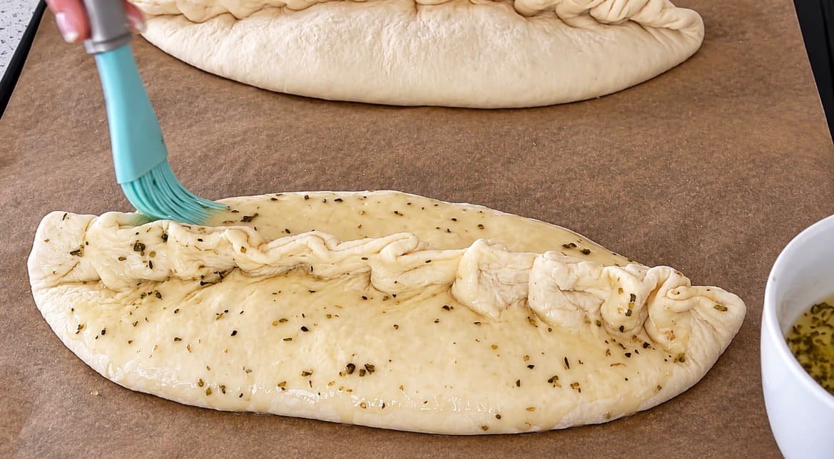 Innbakt pizza pensles med olivenolje og oregano før steking for ekstra smak og en gyllen overflate.